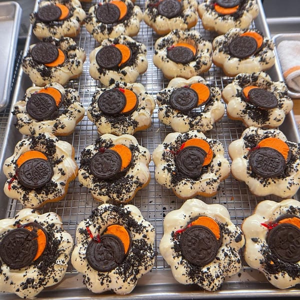 Rows of white-glazed Mochinut donuts with orange candy and crushed cookies on a cooling rack.
