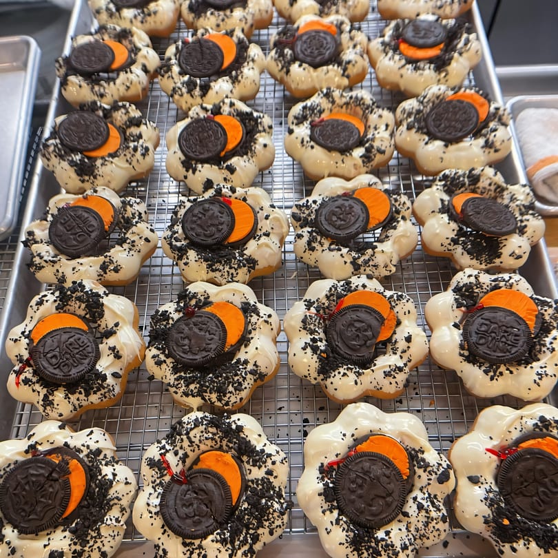 Rows of white-glazed Mochinut donuts with orange and black Oreo toppings on a cooling rack.