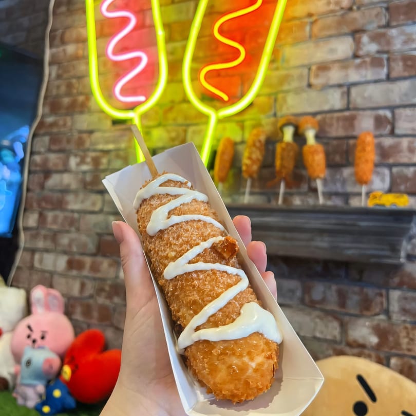 Hand holding a Chung Chun Rice Dog with white sauce, neon hot dog signs in the background.