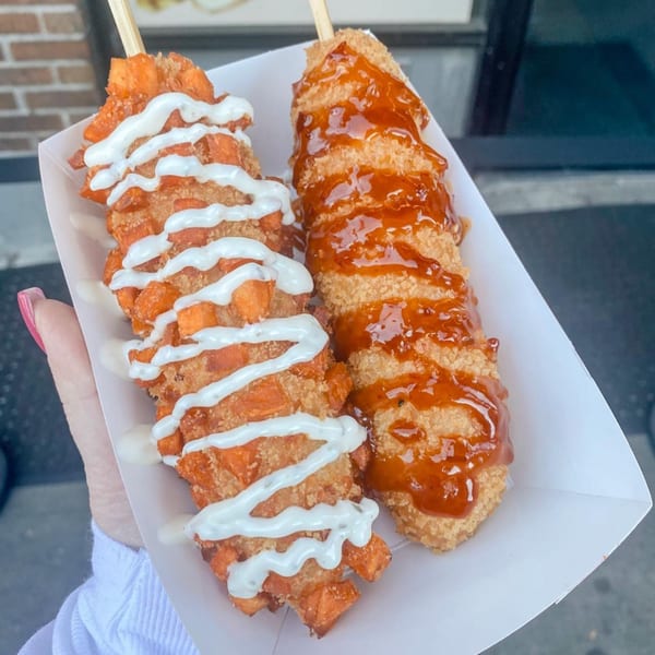 Two Chung Chun Rice Dogs, one with white sauce and the other with brown sauce, held in a white tray.