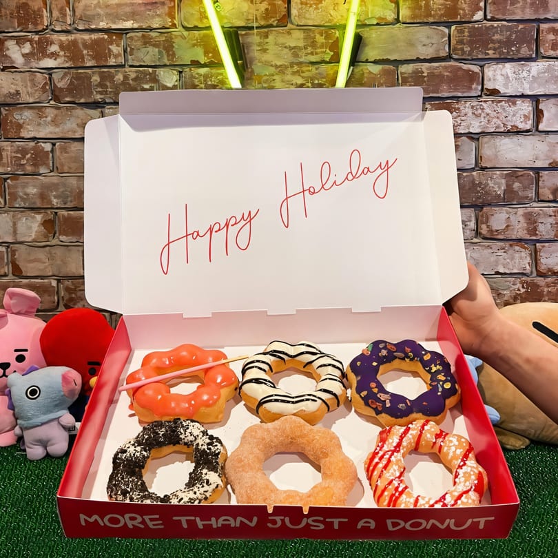 A box of six colorful Mochinut donuts with various toppings, against a brick wall and green turf.