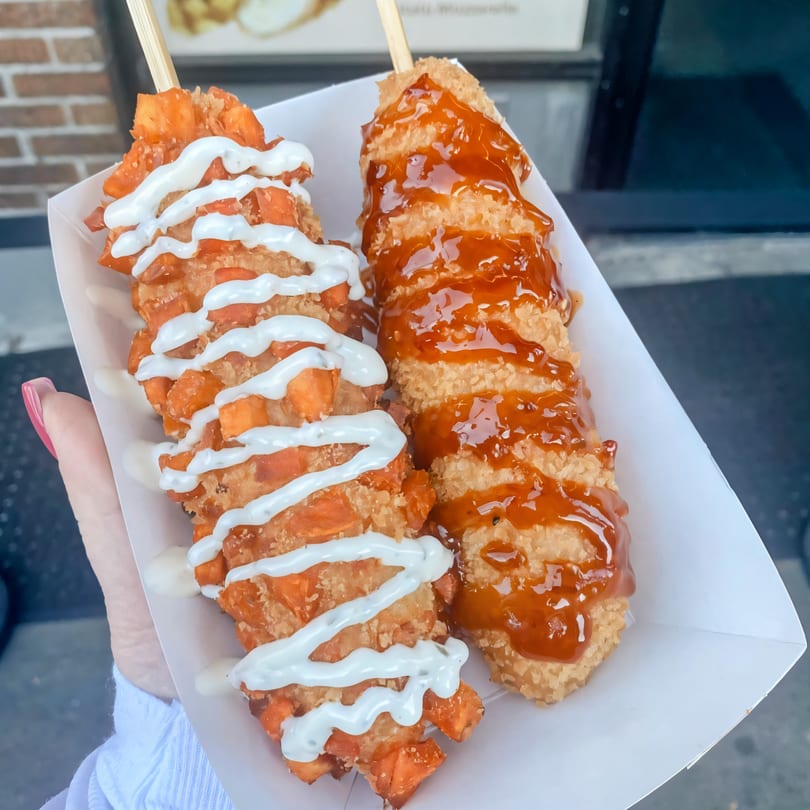 Two Chung Chun Rice Dogs, one with sweet potato and white sauce, the other with brown sauce.